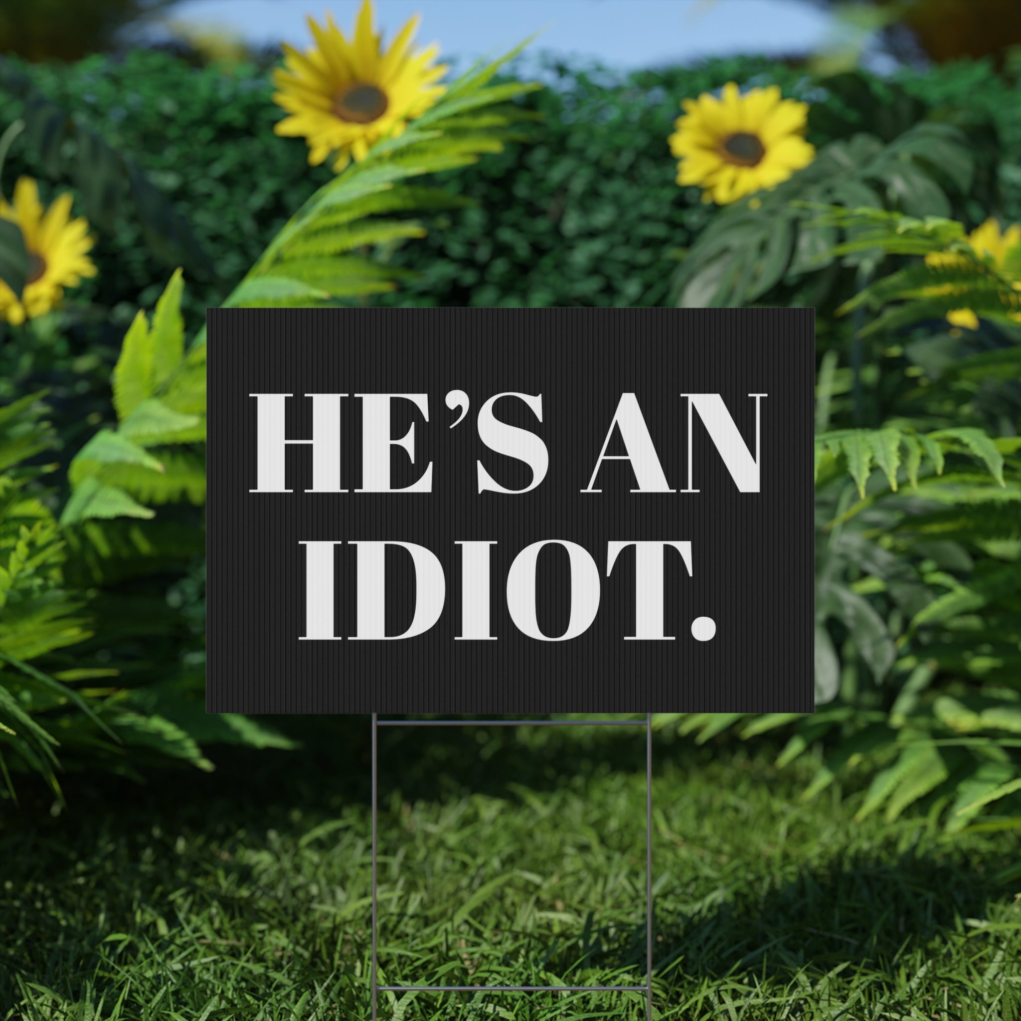 Black sign with white text 'HE'S AN IDIOT.' in a garden setting with sunflowers.