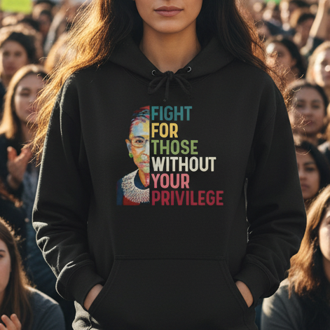 Woman in a black hoodie with a message stands in front of a large crowd at a protest rally.